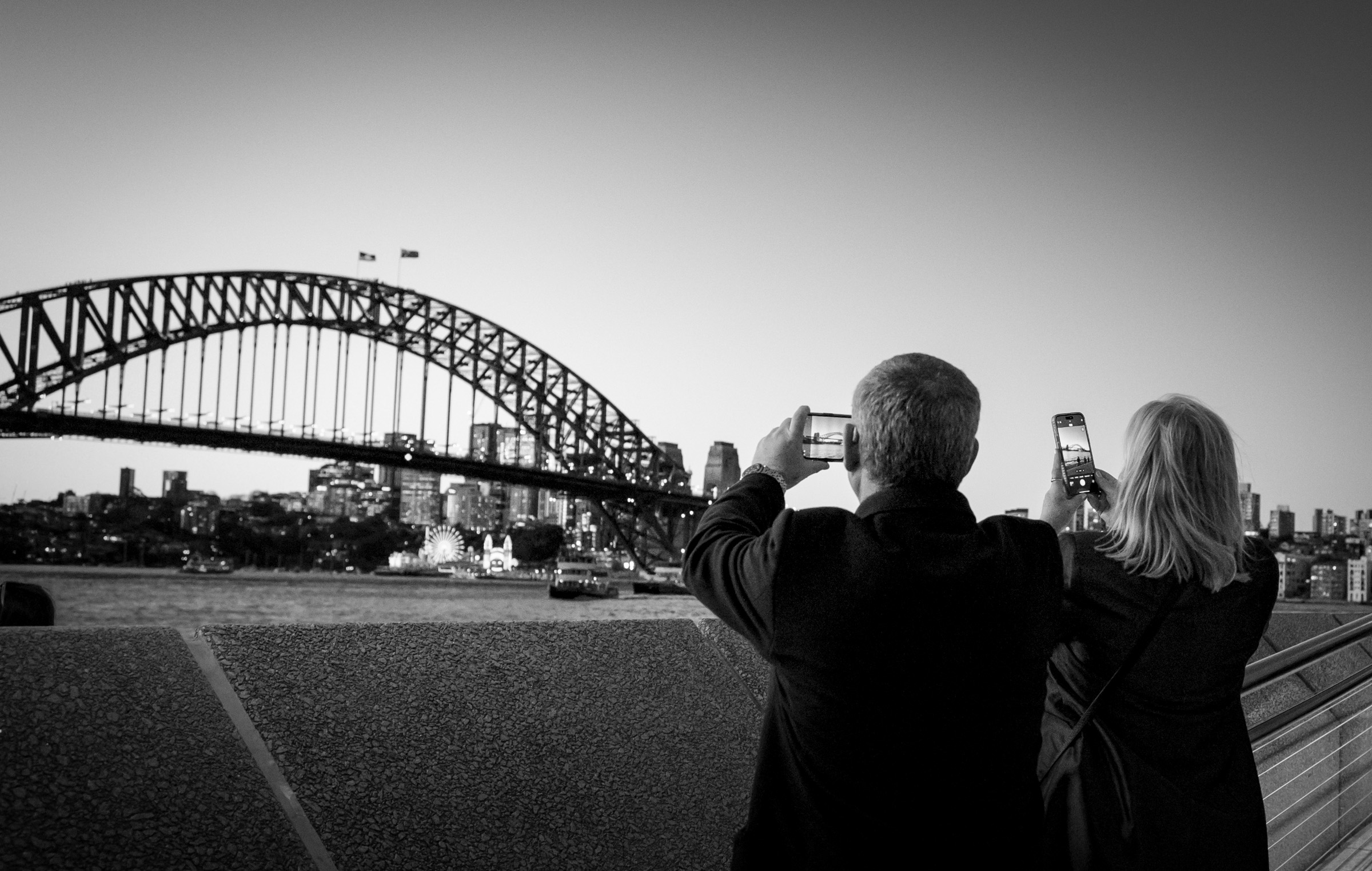 A man and a woman photographing the Sydney Harbour Bridge on their phones, seen from behind