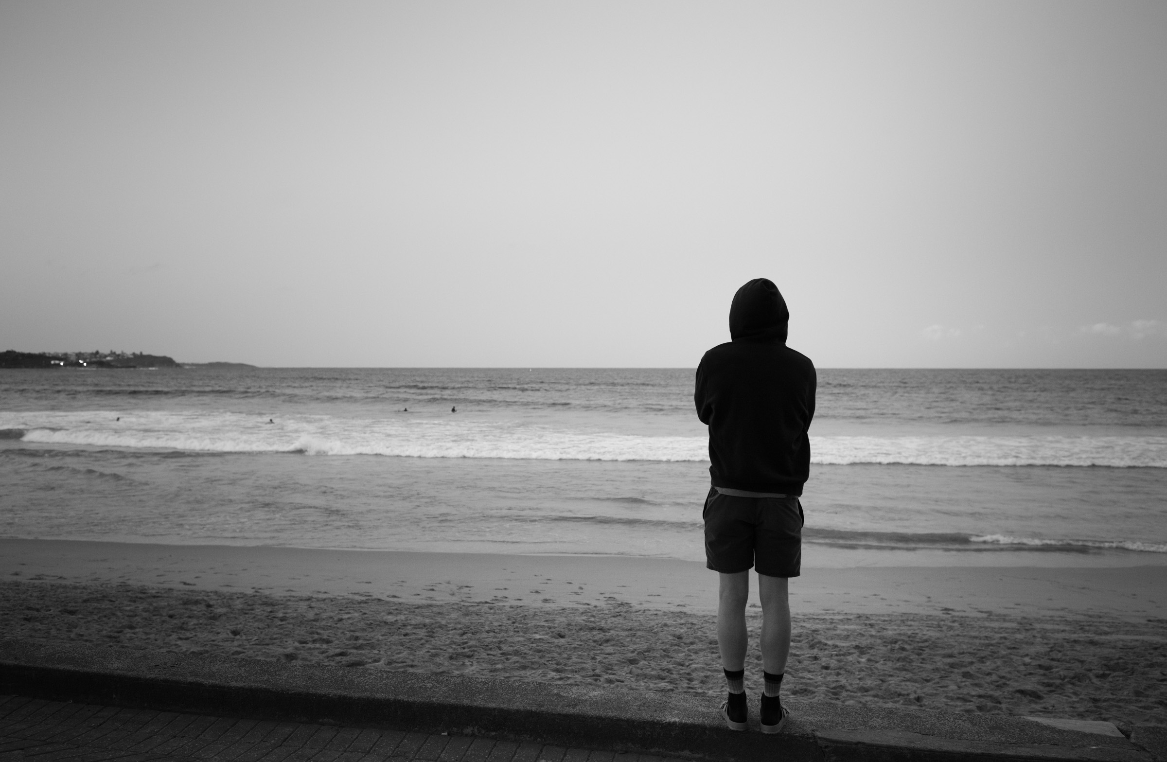 A hooded figure standing alone, facing the ocean, back to camera